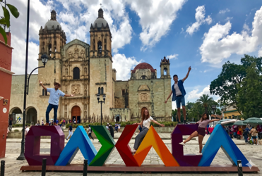Jóvenes posando de forma divertida sobre las letras coloridas de Oaxaca frente al Templo de Santo Domingo de Guzmán bajo un cielo azul.